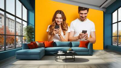 Photo of beautiful people man and woman smiling while both using mobile phones, isolated over yellow background Wall mural