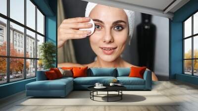 Photo of beautiful woman with towel on head cleaning her face and removing makeup with cotton pad, while standing in bathroom Wall mural