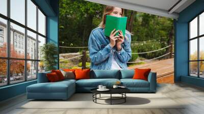 Photo of blonde exciting woman reading book while sitting on bench in green park Wall mural