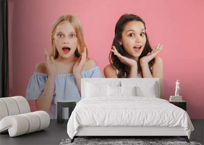 Photo of brunette and blonde princess girls wearing dresses expressing surprise while raising hands at face with open mouth, isolated over pink background Wall mural