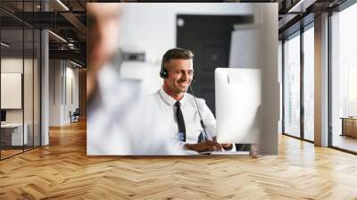 Photo of businesslike man 30s wearing office clothes and headset, smiling while sitting by computer in call center Wall mural