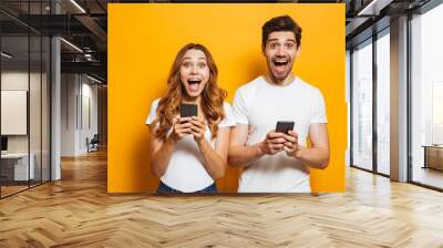 Photo of caucasian beautiful people man and woman screaming, while both using smartphones, isolated over yellow background Wall mural