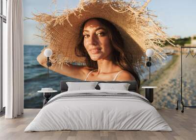 Photo of caucasian brunette woman in swimsuit and straw hat smiling and sunbathing while walking by seaside Wall mural