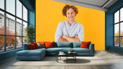 Photo of caucasian cheerful guy 20s with curly hair laughing and standing with arms folded, isolated over yellow background Wall mural