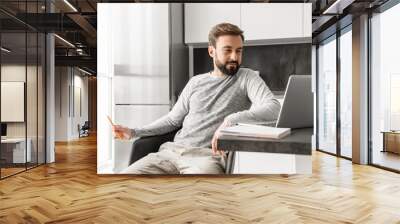 Photo of caucasian man 30s in casual clothing looking on laptop, and working with documents in living room Wall mural