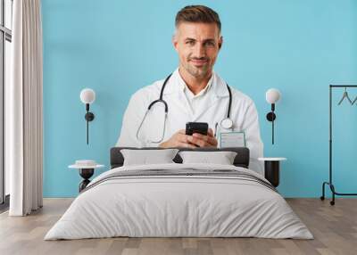 Photo of caucasian medical doctor wearing white coat and stethoscope holding smartphone, standing isolated over blue background Wall mural