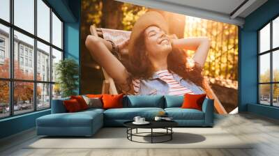 Photo of caucasian relaxed woman 18-20 wearing straw hat, resting with closed eyes and smile in park on sunny day while sitting in lounge chair Wall mural