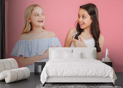 Photo of cheerful little girls 8-10 years old wearing dresses smiling and pointing finger at each other, isolated over pink background Wall mural