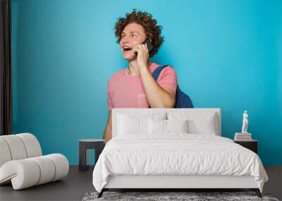 Photo of college guy with curly hair wearing casual clothing and backpack smiling and talking on smartphone, isolated over blue background Wall mural