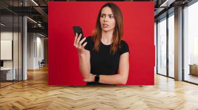 Photo of dissatisfied woman in black clothing looking on mobile phone with confusion or misunderstanding, over red background Wall mural