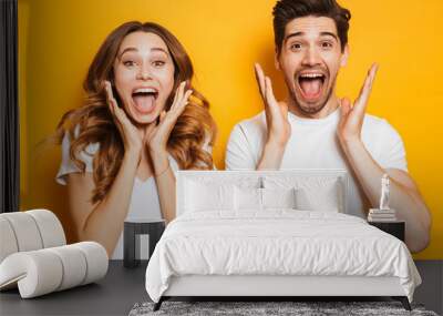 Photo of emotional couple man and woman in basic clothing screaming in surprise or delight and raising arms, isolated over yellow background Wall mural