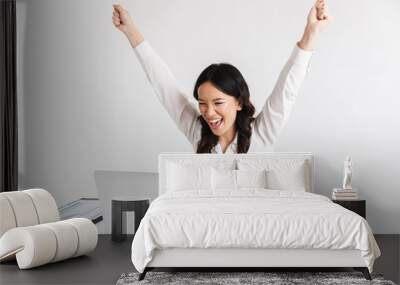 Photo of excited chinese businesswoman with long dark hair screaming with raised arms while working with documents and laptop, isolated over white background in studio Wall mural