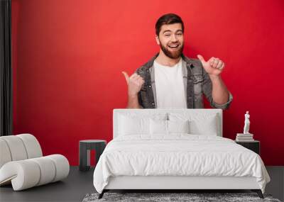 Photo of handsome man 30s in casual clothing gesturing fingers aside on copyspace with surprise and smile, isolated over red background Wall mural