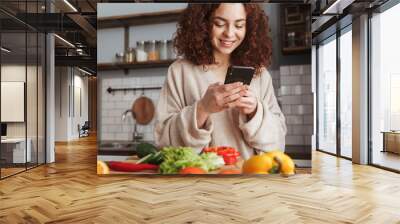 Photo of happy caucasian woman holding smartphone while cooking salad with fresh vegetables at home Wall mural