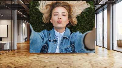 Photo of happy kind woman taking selfie photo and sending kiss while lying green grass in park Wall mural