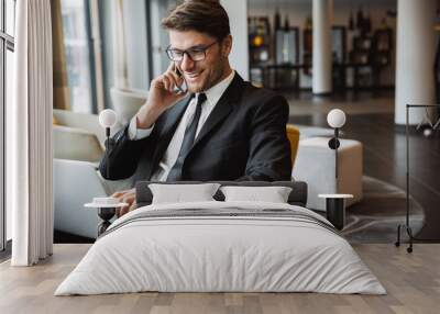 Photo of happy young businessman using laptop computer and talking on smartphone in hotel hall Wall mural