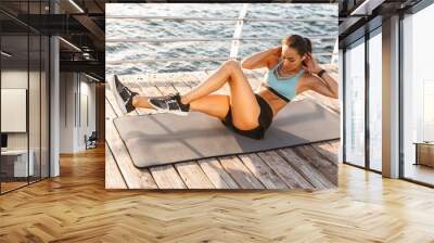 Photo of healthy fitness woman 20s in sportswear lying on exercise mat near seaside, and doing crunches with arms behind head Wall mural