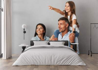 Photo of joyful family woman and man smiling and looking aside while pretty girl sitting on the neck of her happy father, isolated over gray background Wall mural