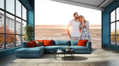 Photo of joyful young couple smiling and hugging while walking on sunny beach Wall mural