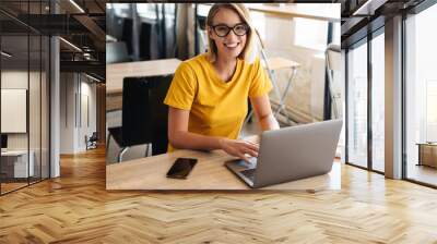 Photo of joyful young woman using laptop and smiling while sitting Wall mural