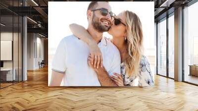 Photo of lovely young woman kissing and hugging handsome man while walking on sunny beach Wall mural