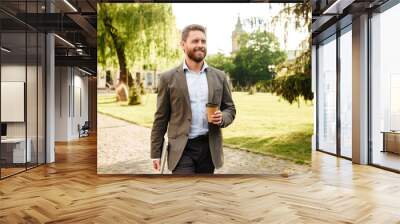 Photo of mature joyous man in gray classical suit, walking along park carrying takeaway coffee and silver laptop Wall mural