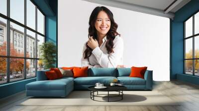 Photo of optimistic asian woman with long dark hair looking aside at copyspace and laughing, isolated over white background in studio Wall mural