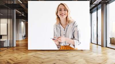 Photo of optimistic beautiful woman wearing glasses smiling at camera and typing on smartphone Wall mural