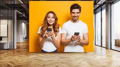 Photo of pleased attractive people man and woman smiling, while both using mobile phones, isolated over yellow background Wall mural