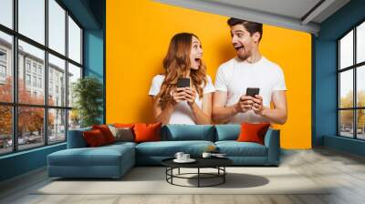 Photo of positive excited people man and woman screaming and looking at each other while both using mobile phones, isolated over yellow background Wall mural