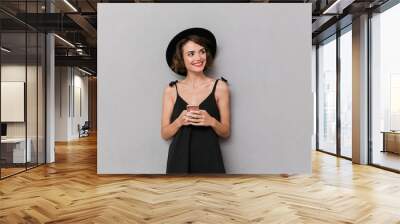 photo of positive woman 20s wearing black dress and hat smiling at camera while holding smartphone,  Wall mural