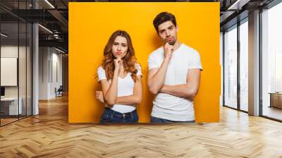 Photo of resented man and woman in casual clothes standing together and touching chin with irritated look, isolated over yellow background Wall mural