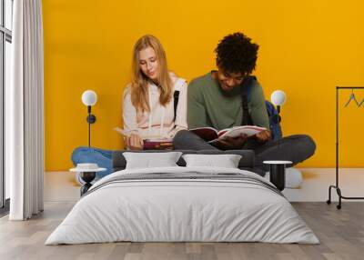 Photo of smart students 16-18 using reading books while sitting on floor with legs crossed, isolated over yellow background Wall mural