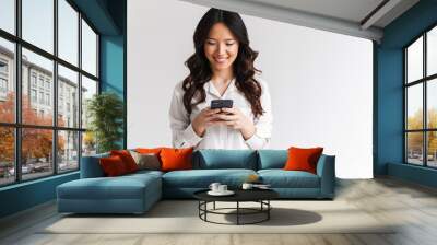 Photo of smiling asian woman with long dark hair holding and using black mobile phone, isolated over white background in studio Wall mural