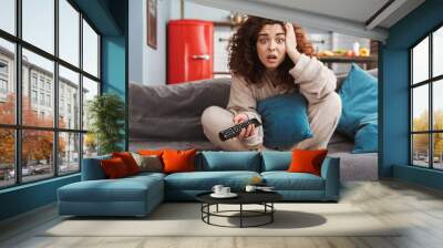 Photo of stressed young woman sitting on sofa in apartment and holding remote control Wall mural