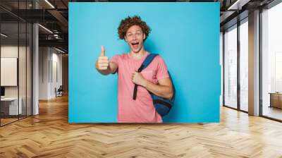 Photo of student guy with curly hair wearing casual clothing and backpack smiling and showing thumb up, isolated over blue background Wall mural