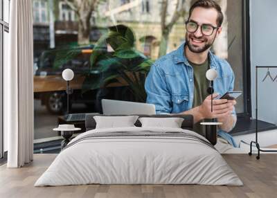 Photo of successful young man holding smartphone while working on laptop in city cafe outdoors Wall mural