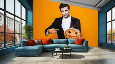 Photo of terrifying dead man on halloween wearing classical suit and creepy makeup holding carved pumpkin, isolated over yellow background Wall mural