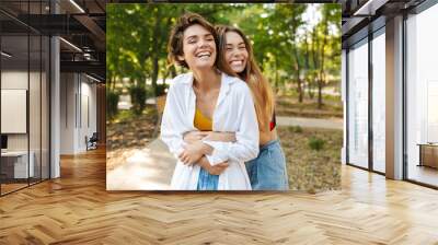 Photo of two women hugging and laughing while walking in green park Wall mural