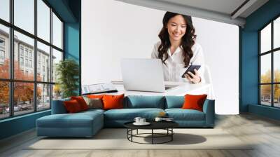 Photo of young asian businesswoman with long dark hair sitting at table and holding smartphone while working with laptop, isolated over white background in studio Wall mural