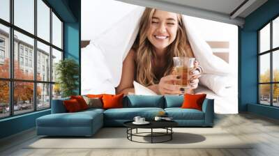 Photo of young laughing woman reading book and drinking tea while lying Wall mural