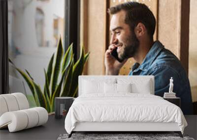 Photo of young smiling man talking on cellphone with laptop and clipboard while working in cafe indoors Wall mural