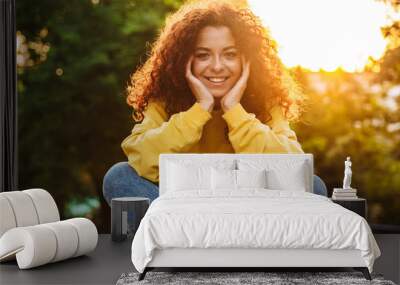 Pleased happy cute young student curly girl sitting on bench outdoors in nature park with beautiful sunlight. Wall mural