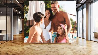 Portrait closeup of joyful european man and woman with kids smiling, while resting near luxury swimming pool with deckchairs and umbrellas during vacation Wall mural