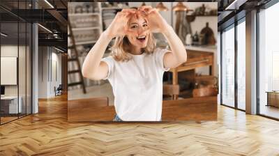 Portrait of adorable blond girl 20s wearing casual t-shirt smiling and showing heart shape with fingers while standing in stylish wooden kitchen at home Wall mural