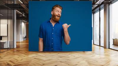 Portrait of adult handsome stylish redhead bearded surprised man in blue shirt with opened mouth,looking and pointing aside with thumb finger , while standing over isolated blue background Wall mural