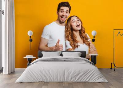 Portrait of cheerful people man and woman in basic clothing smiling and showing thumb up at camera, isolated over yellow background Wall mural