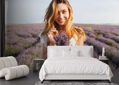 Portrait of cheerful pretty woman in dress holding bunch of flowers, while walking outdoor through lavender field in summer Wall mural