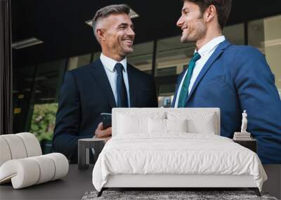 Portrait of happy businessmen partners standing outside job center and using cellphone together during working meeting Wall mural