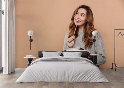 Portrait of happy thinking female student in warm sweater holding pencil and notebooks, looking aside Wall mural